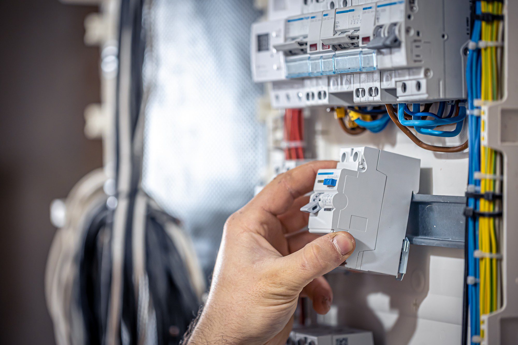 This image shows an electrical installation in progress. A person's hand is holding a device which appears to be a circuit breaker or some kind of electrical protection device, ready to be installed in the electrical panel. The panel has several other breakers already in place, and there are wires connected to these breakers that ensure electricity distribution throughout a building. This is typically part of the electrical infrastructure in residential or commercial buildings. The wiring is neatly organized with cable ties and trunking for protection and structure. The scene reflects electrical maintenance work or setup, usually carried out by a qualified electrician.