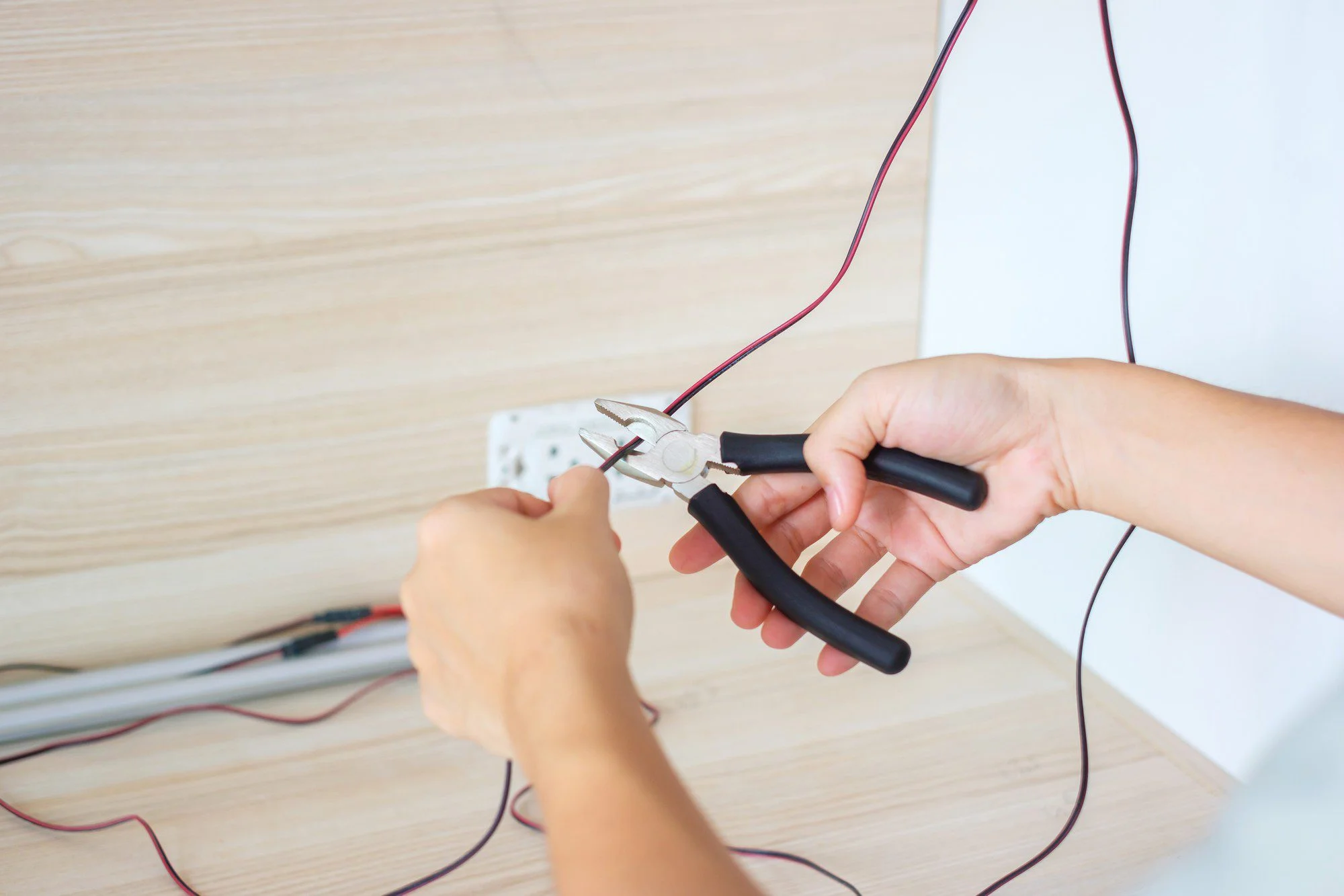 The image shows a person's hands working on an electrical socket. They are holding a pair of pliers and manipulating wires that are connected to the back of the electrical outlet. The wires appear to be red and black, indicating they are likely part of a low-voltage DC circuit or possibly the live (hot) and neutral wires of an AC circuit, but this latter point would be unusual given the colour coding standards for AC wiring in many countries. The person appears to be in the midst of installing or repairing the outlet. The setting looks like a domestic or office environment, based upon the wooden surface and the relaxed background. Safety is important when working with electricity, and it is critical to ensure that the circuit is de-energised before touching any wires.