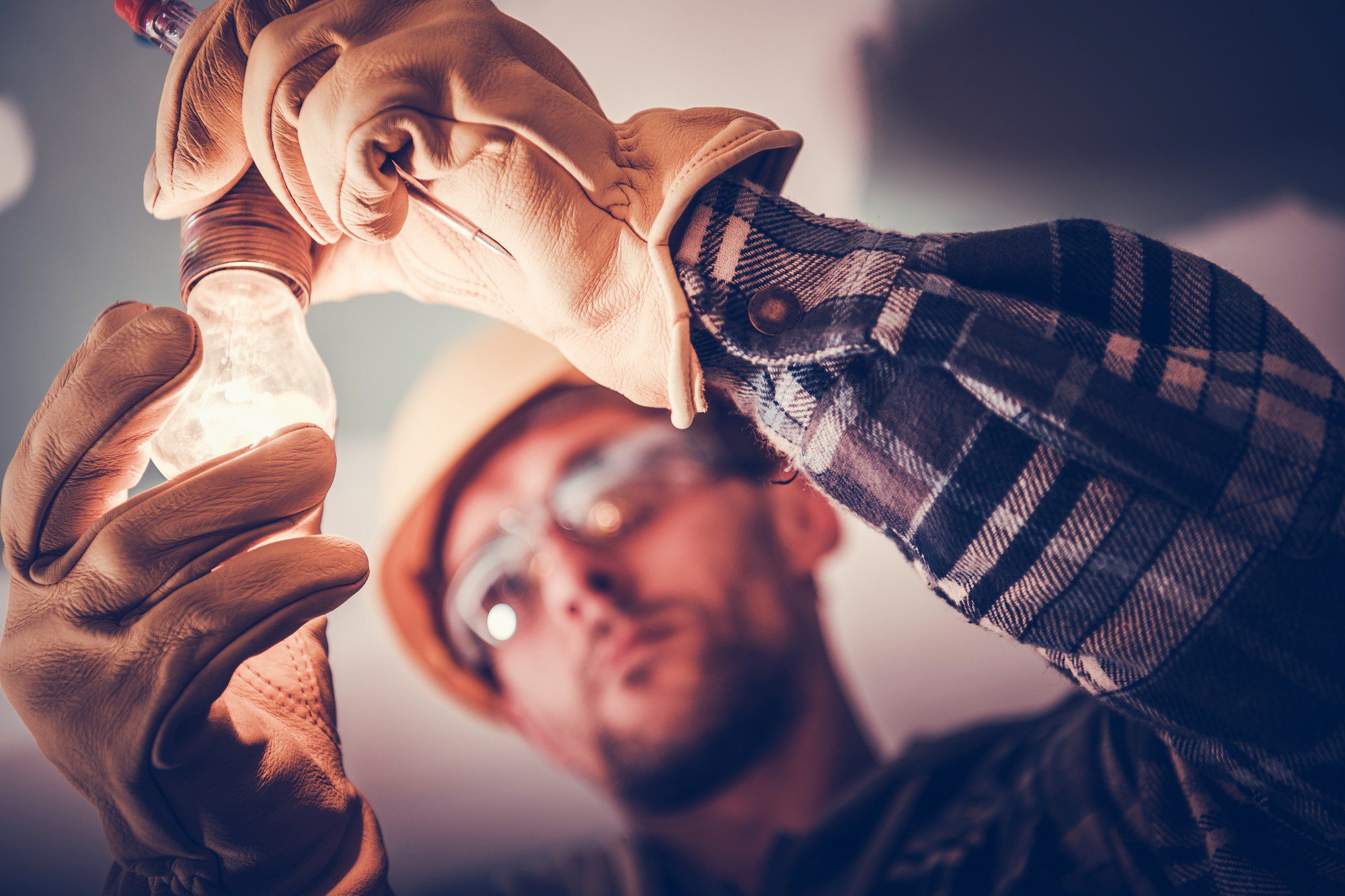 The image shows a person wearing a hard hat and safety goggles, focused on screwing in a light bulb. The individual appears to be following safety protocols, as indicated by the protective equipment. The light bulb is illuminated, suggesting that the electrical circuit is active, and highlights the importance of safety when dealing with electrical components. The person is also wearing gloves, which could be for additional protection or for better grip. The photograph has a warm tone and a shallow depth of field, focusing on the hands and the light bulb, while the background is blurred.