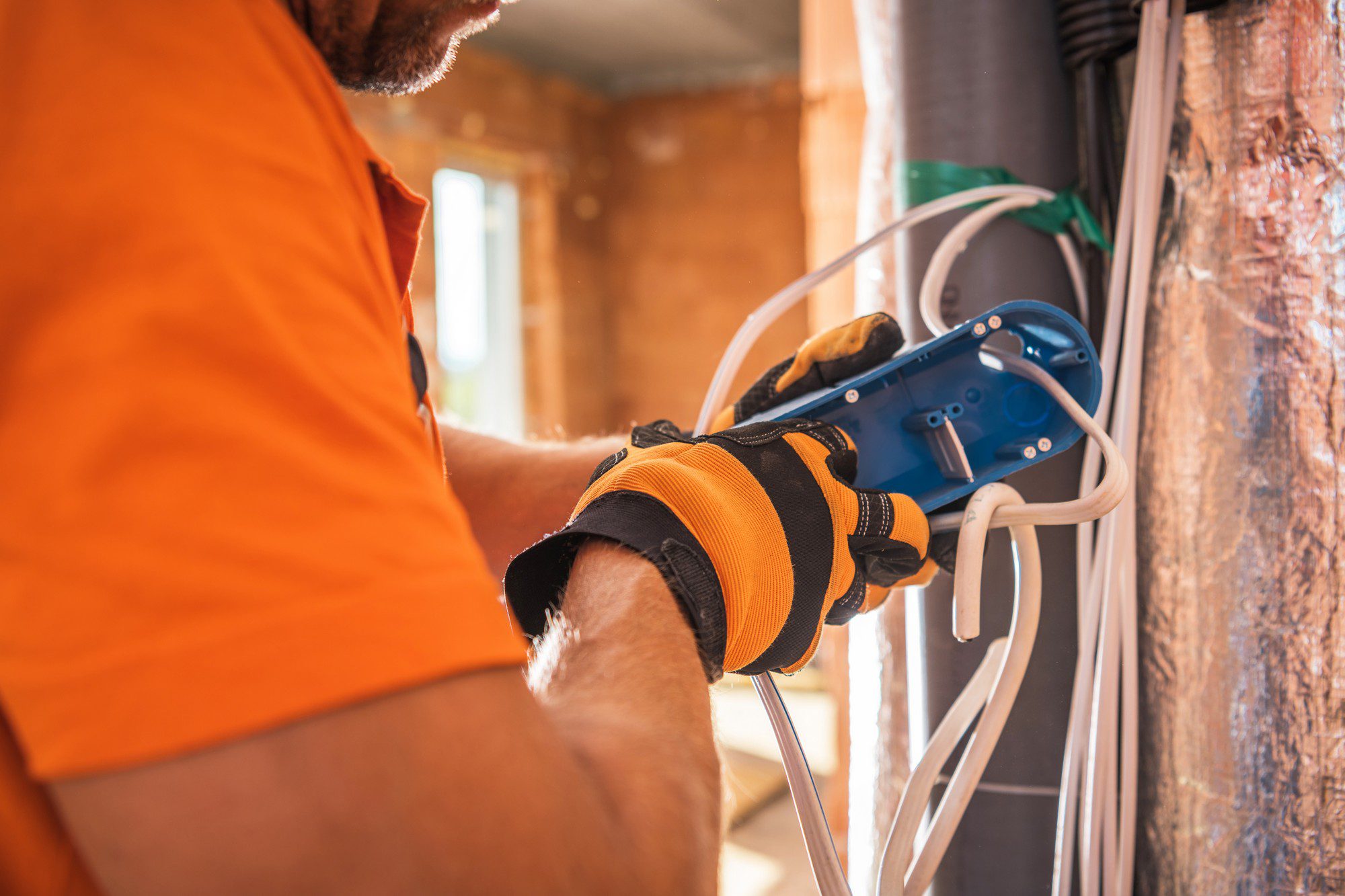 In this image, we see a close-up of a person's arm and hand as they are working on what appears to be an electrical installation. The person is wearing an orange shirt and work gloves. They are holding a blue electrical box or junction box against a wall and routing several white electrical cables into it. The wall shows bare wood framing, suggesting that the work is taking place in a building under construction or undergoing renovation. The presence of insulation material can also be seen in the wall, which indicates that energy efficiency measures are being considered. The focus is on the task and the hands, which are centrally located in the image. The person's head and most of their body are out of frame. The details visible suggest that this is an environment where electrical or construction work is being performed, and safety measures such as gloves are being observed.