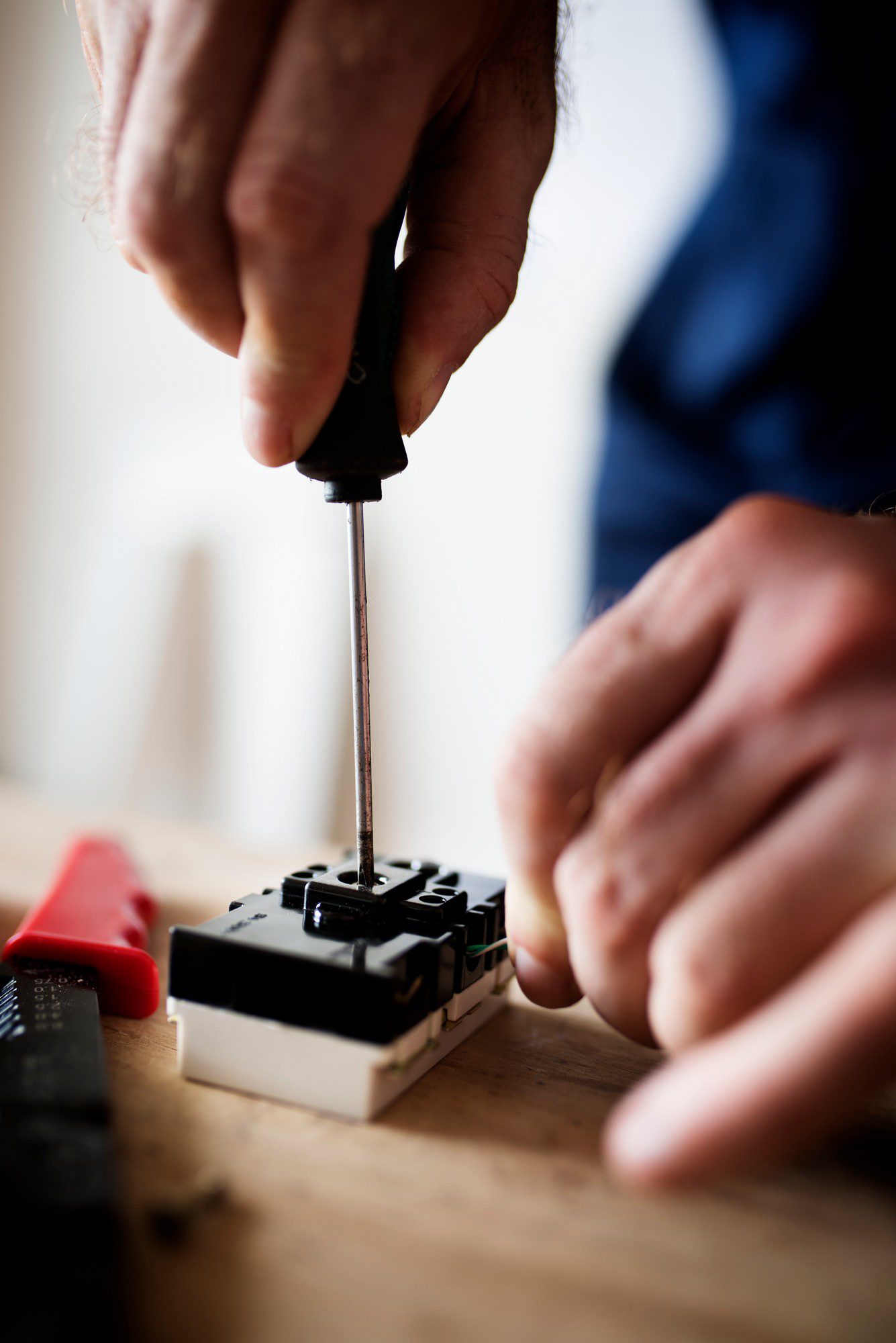 The image shows a close-up of a person's hands working with a screwdriver on an electrical component. You can see the screwdriver being used to either tighten or loosen a screw on the piece of equipment, which appears to be mounted on a workbench or table. The focus is on the hands and the screwdriver, with a blurred background suggesting a workshop setting. In the foreground, there are out-of-focus tools or parts, possibly related to the work being performed.