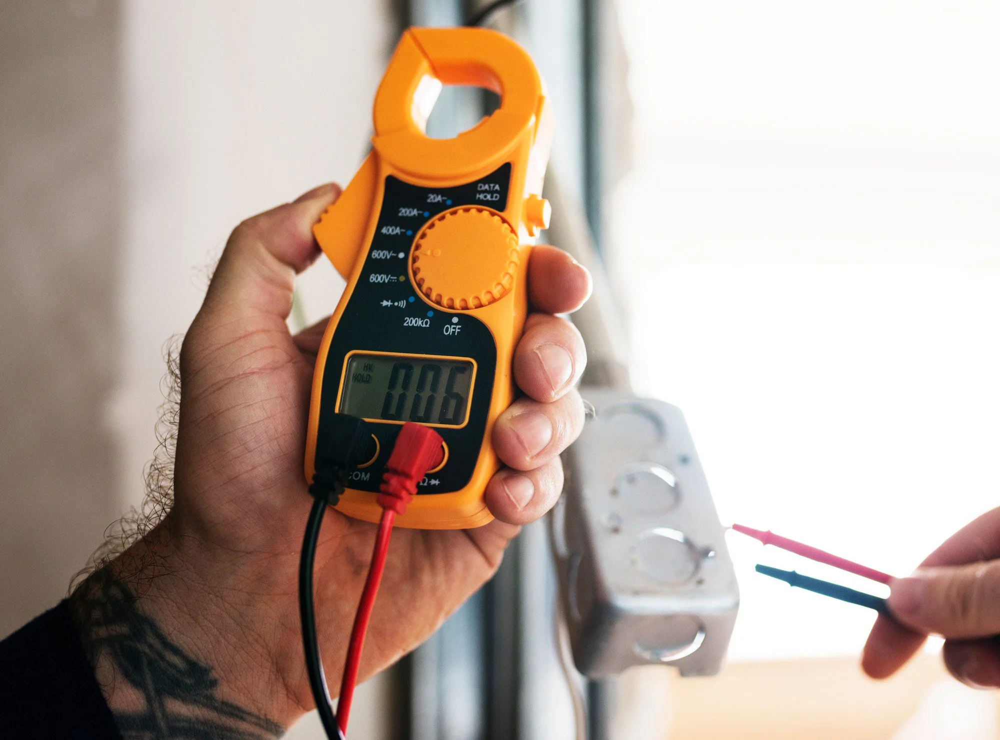 This image shows a person holding a digital clamp meter, which is an electrical test tool that combines a basic digital multimeter with a current sensor. The clamp meter is being used near what appears to be an electrical junction box or installation. On the display of the clamp meter, you can see a reading of "0.06," possibly indicating a measurement of current, voltage, or another electrical parameter, depending on the setting of the device. The person's other hand is holding the meter's test leads, which are colour-coded red and black, a standard in electrical work to denote positive and negative leads, respectively. The environment suggests the individual is performing an electrical testing or troubleshooting task.
