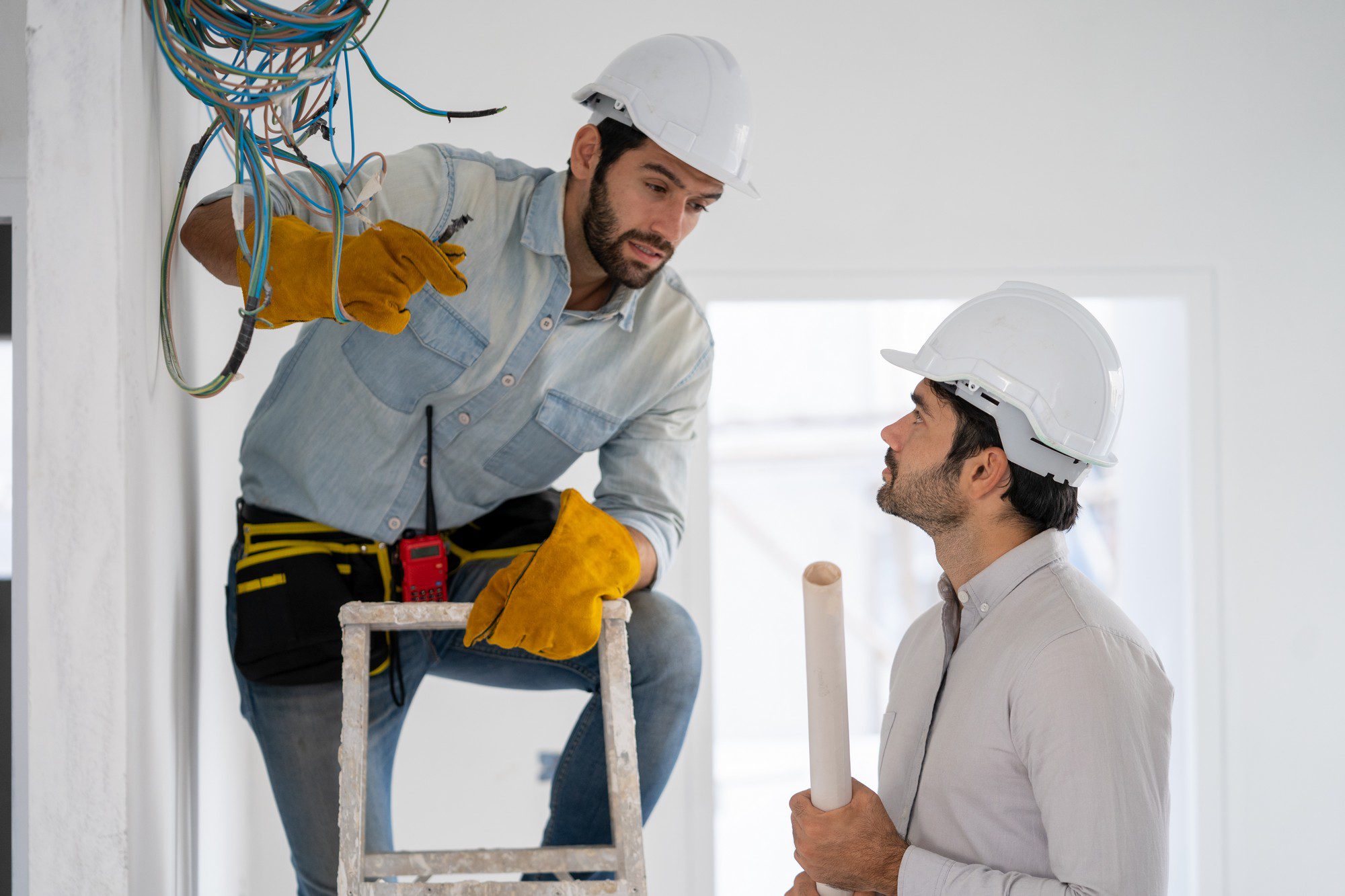 The image shows two people who appear to be working on an electrical installation in a building or home under construction or renovation. The individual on the left is wearing a blue button-up shirt with rolled sleeves, yellow work gloves, a tool belt, and a white safety helmet. They are standing on a ladder and appear to be handling electrical cables that emerge from a conduit in the wall, indicating they might be an electrician.The person on the right is wearing a lighter-coloured long-sleeved shirt and also has a white safety helmet. They are holding a long white tube or conduit, possibly discussing with the electrician where it should go or how to install it. Both individuals seem to be engaged in a work-related conversation or planning.The surroundings suggest a work-in-progress environment with white walls and what seems to be a partially completed electrical system. Safety seems to be a priority, as they are both wearing helmets, and the one handling the wiring is wearing protective gloves.