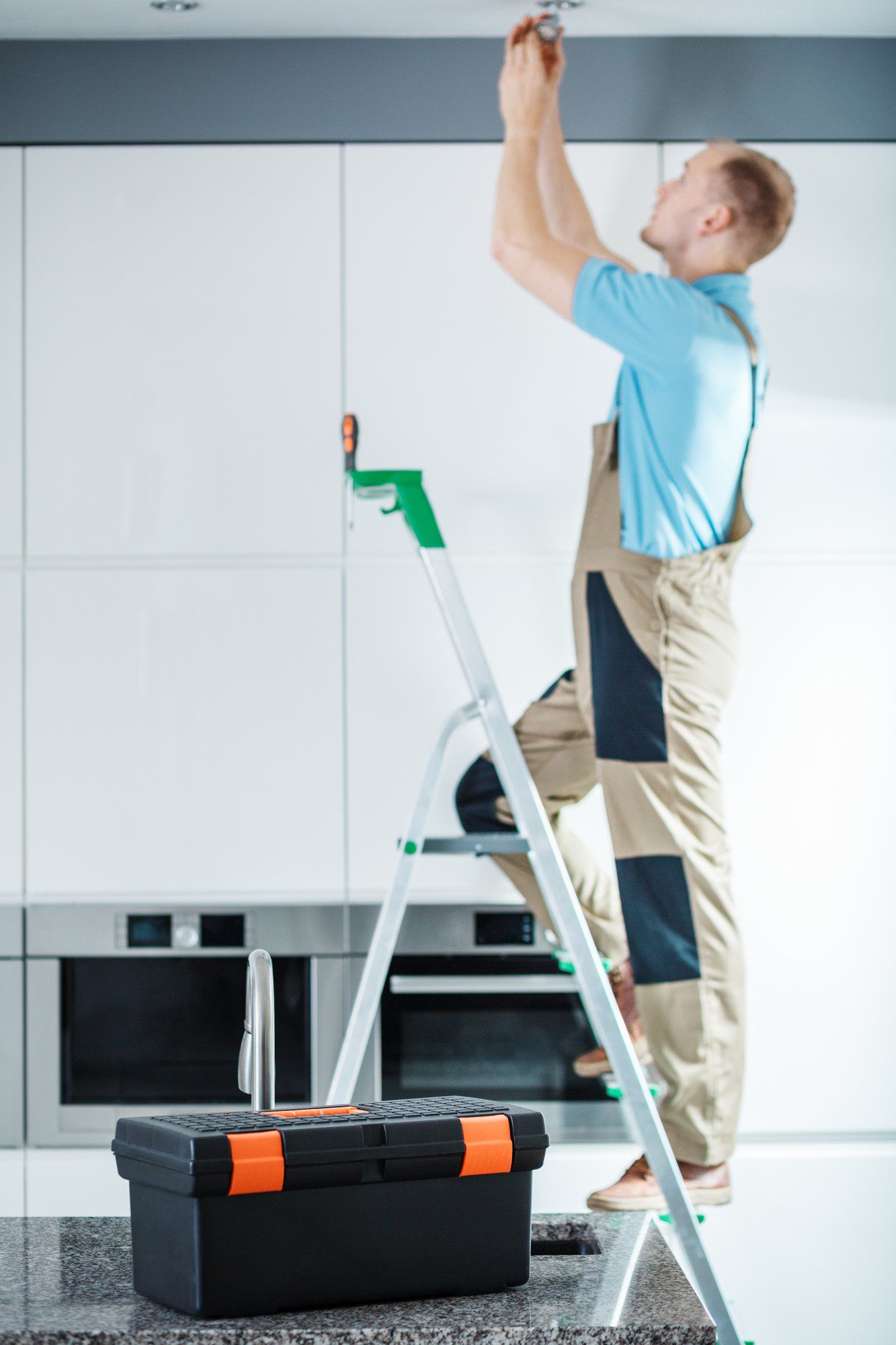 The image shows a person standing on a step ladder, reaching up towards the ceiling, possibly fixing something or installing a device. They are wearing a blue shirt and khaki pants, which suggests they could be a repair person or a DIY enthusiast working on a home improvement project. In the foreground, there is a black and orange toolbox placed on a kitchen countertop, indicating that the person has tools at their disposal for the task. The setting appears to be a modern kitchen with white cabinetry and built-in appliances.