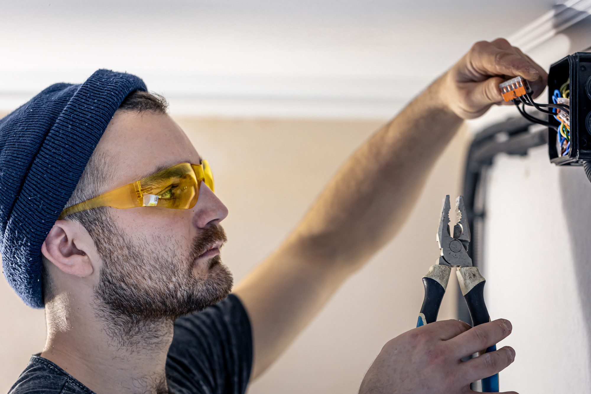 The image shows a person engaged in electrical work. The individual is wearing safety glasses and a beanie hat, and is reaching into an open electrical junction box on the wall, likely working on wiring. The person is holding a pair of pliers, which is a common tool for electricians to twist wires, cut wire insulation, or hold components. Their focus on the task suggests meticulous work, likely to ensure electrical connections are made safely and correctly. Safety practices such as wearing protective eyewear are important in this kind of work to prevent injuries from potential sparks or debris.