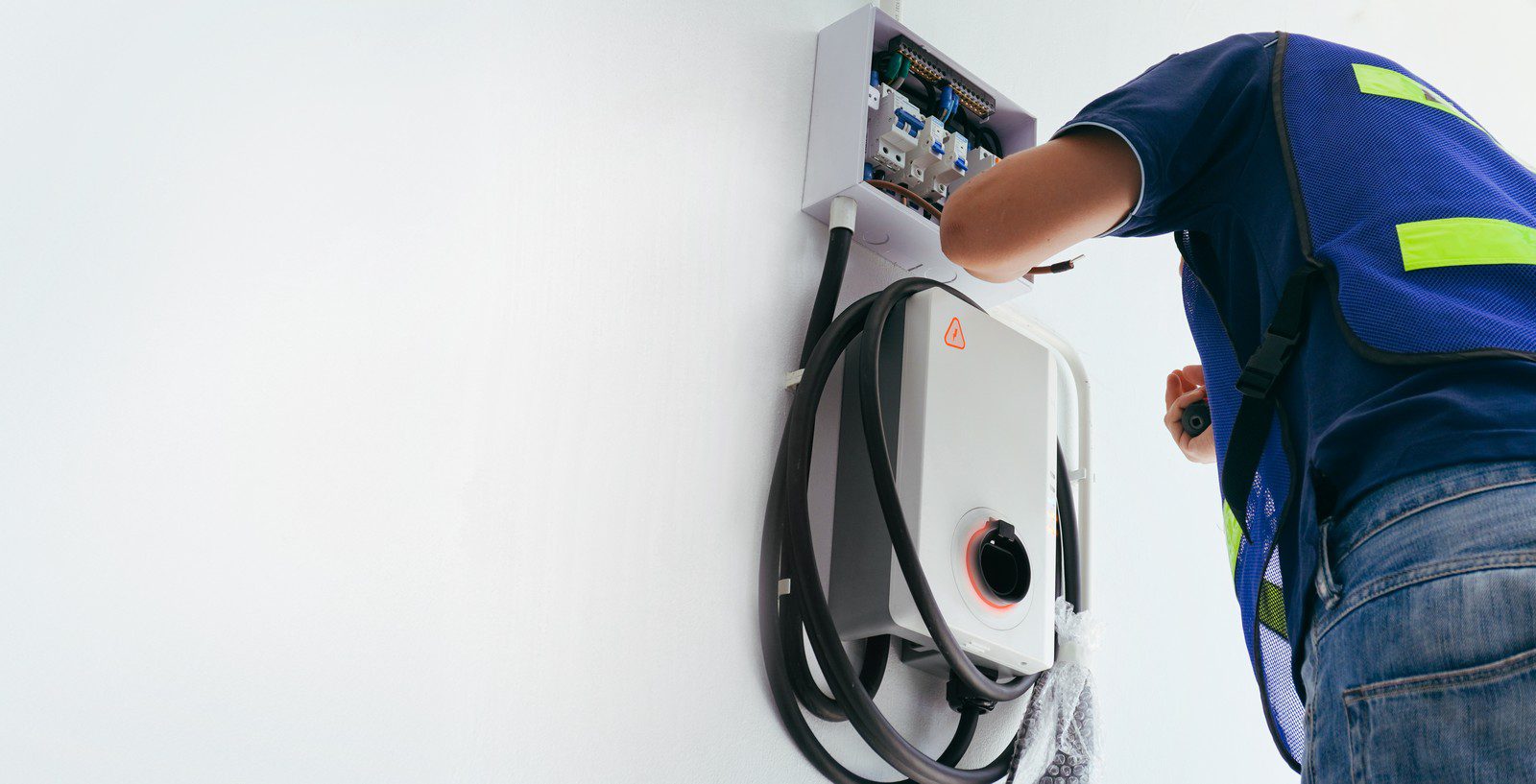 The image shows a person in a blue shirt with reflective safety vest, likely an electrician or technician, who is working on an electrical installation. Specifically, the person is attending to an electric vehicle (EV) charging station that is mounted on a wall. The EV charger has a thick cable coiled around it, and the charging plug is visible at the front. The cover of the charger is removed, revealing the internal components and wiring, and the technician appears to be either installing, inspecting, or repairing the unit. The white background suggests this might be an indoor setting, possibly a garage or a dedicated charging station area.