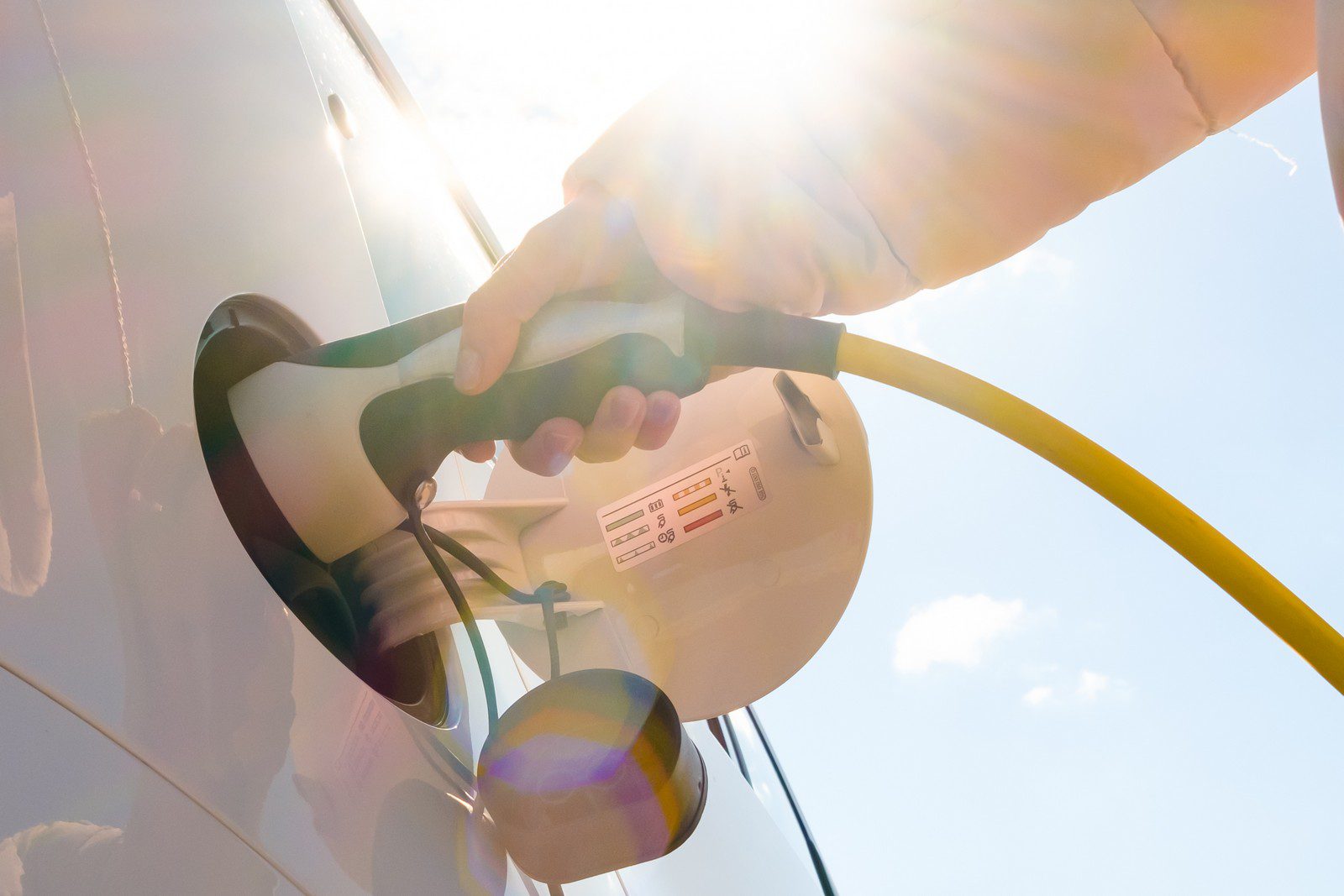 The image shows an electric vehicle (EV) charging station with a charging cable plugged into a car. A person's hand is holding the connector as it inserts or secures it into the charging port of the vehicle. The sunlight is causing some lens flare, which adds a bright, somewhat overexposed effect to the photo.