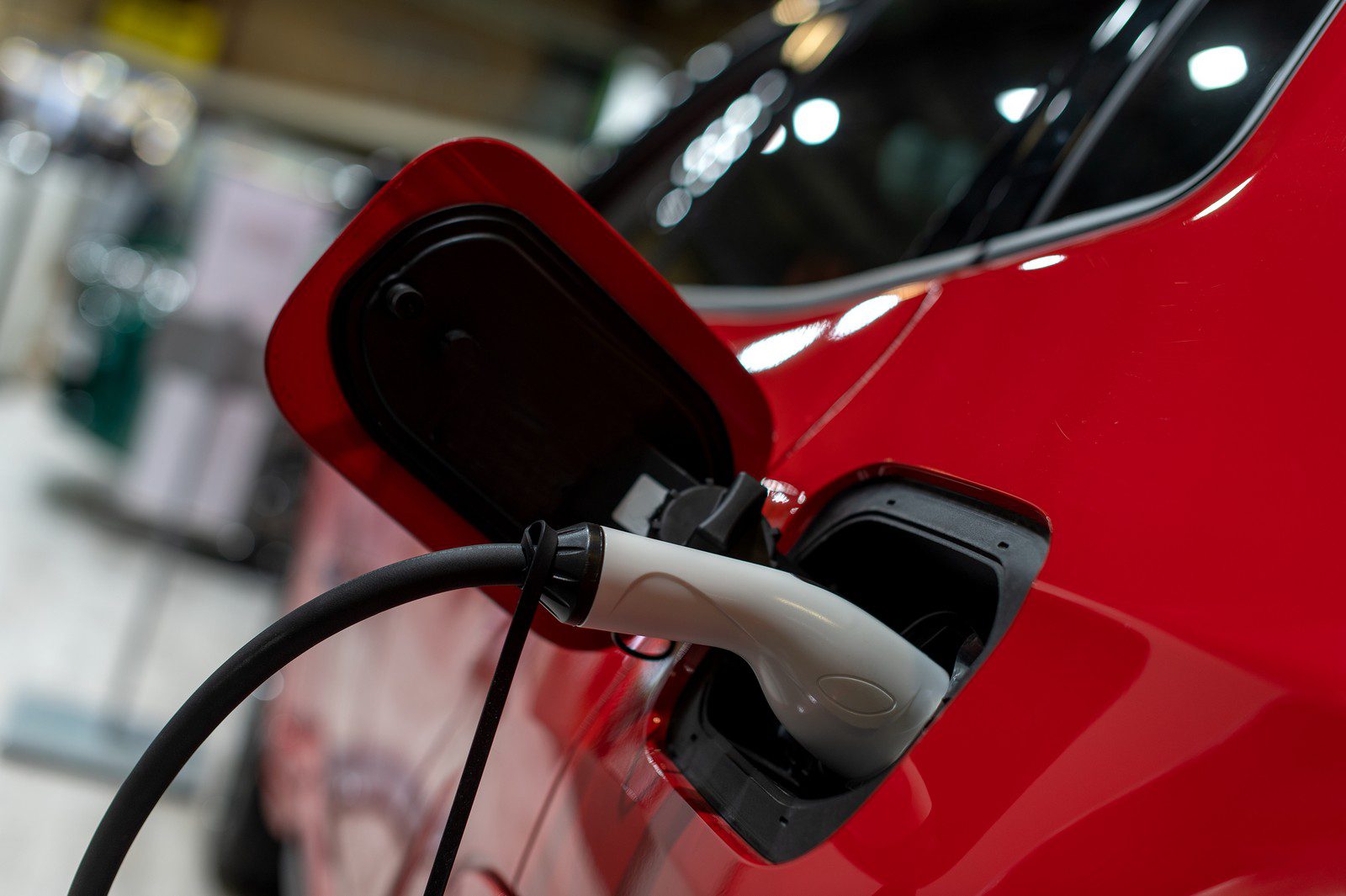 The image shows an electric vehicle (EV) being charged. The charging cable is connected to the charging port of the car, which is often located where a fuel filler would be on a conventional internal combustion engine vehicle. The car has a shiny red exterior, and the charging cable has a white connector which is plugged into the vehicle. This image represents the process of recharging an electric vehicle's battery, which is essential for the operation of EVs.