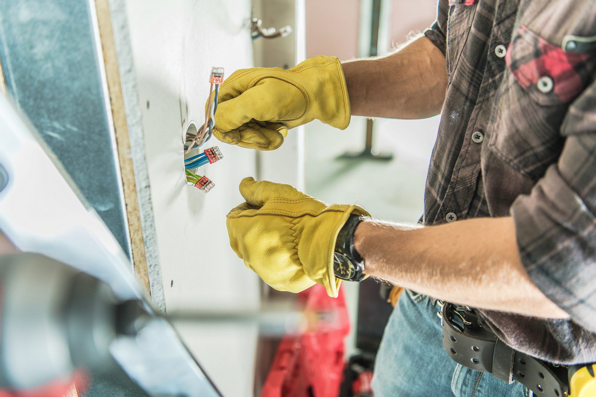 The image shows an individual working with electrical wiring. The person is wearing yellow safety gloves to protect their hands while they handle the electrical components. They are holding some electrical wires that have been capped with wire nuts, indicating that they are either in the process of installing or repairing something related to the building's electrical system. The person is wearing a plaid shirt, has a watch on their wrist, and is equipped with a tool belt around their waist, which suggests they are a professional tradesperson, likely an electrician. The environment indicates an indoor setting, possibly during construction or renovation, as the wall behind the wiring is unfinished.