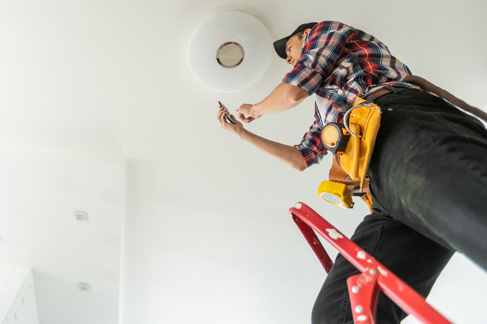 The image shows a person standing on a ladder and working on a ceiling fixture. The individual is wearing a plaid shirt, a baseball cap, and a tool belt with various tools and equipment, suggesting they might be an electrician or a handyman in the process of installing or repairing the fixture. The environment looks like a domestic or office interior with white walls and a bright, clean aesthetic. The angle of the photo is from below, focusing on the worker and the task at hand while providing an upwards perspective.