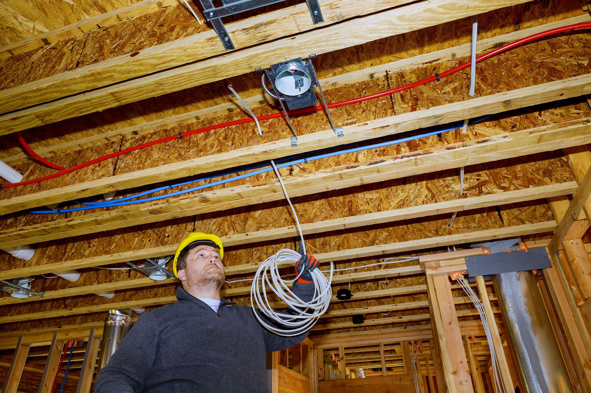 This image shows the interior of a building under construction with a focus on the ceiling area where electrical and possibly plumbing installation work is in progress. At the centre of the photo, there is a person who appears to be an electrician or a construction worker, wearing a hard hat and holding a bundle of electrical cables. The worker is looking upwards at the ceiling work.The ceiling is unfinished, with exposed wooden beams and OSB (oriented strand board) sheathing. You can see multiple utilities running across the ceiling joists, including red and blue PEX (cross-linked polyethylene) pipes, which are often used for plumbing, and various conduits and electrical boxes for the electrical systems.On the right side, there's a ductwork system, likely for heating, ventilation, or air conditioning (HVAC), and recessed light fixtures installed between the joists. The setting indicates that the infrastructure for the building's essential services is being put in place before the walls and ceilings are closed with drywall or other finishing materials.