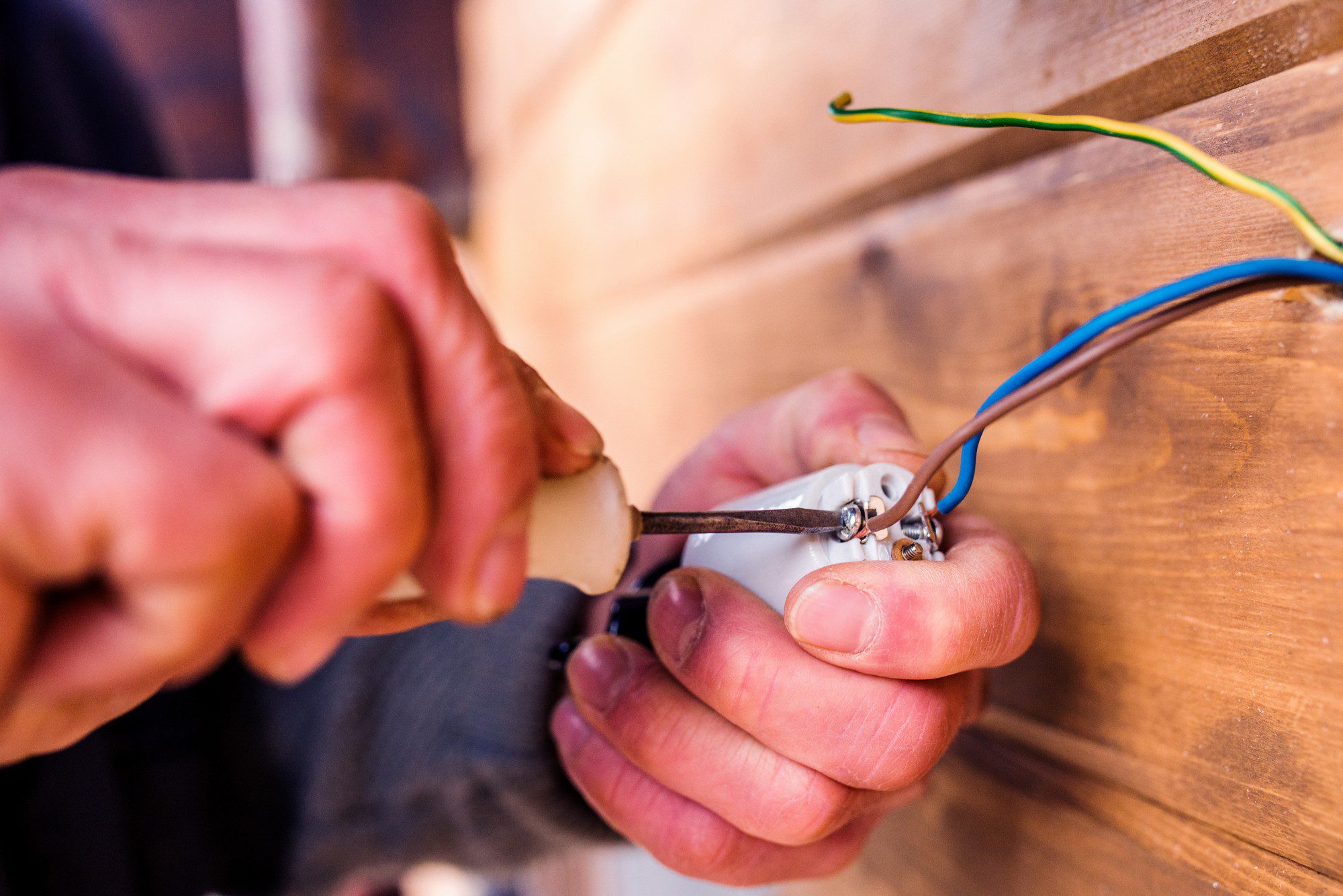 This image shows a person's hands working with electrical wires and a tool. The individual appears to be in the process of stripping the insulation from the ends of the wires, which come in blue, green, and yellow colours, indicating they are likely conducting electrical work, possibly preparing to connect the wires to a terminal or switch. The hands are holding a pair of pliers, and it seems the person is twisting or manipulating the wire with the tool. The background is somewhat blurred but looks like wooden panels, suggesting the work might be taking place in a construction or renovation setting.