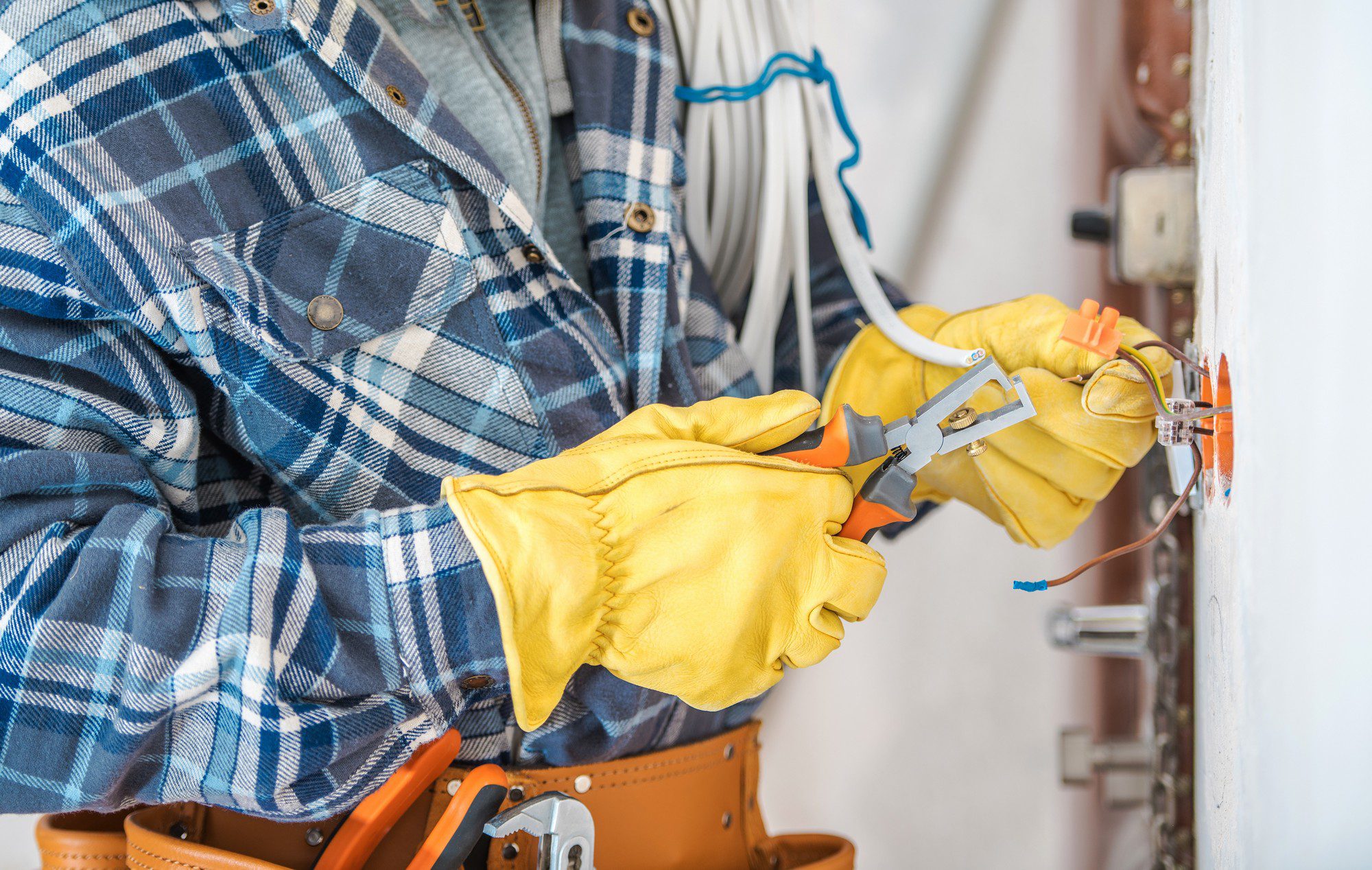 This image shows a person wearing a plaid shirt and yellow work gloves manipulating electrical wires, suggesting electrical work or installation. The individual is using pliers to work on a wiring connection within an open junction box mounted on the wall. Safety precautions appear to have been taken, such as using insulated tools and wearing gloves to prevent electrical shock. The image also displays a partially visible tool belt, indicating that the person is likely a professional electrician or has experience with electrical work.