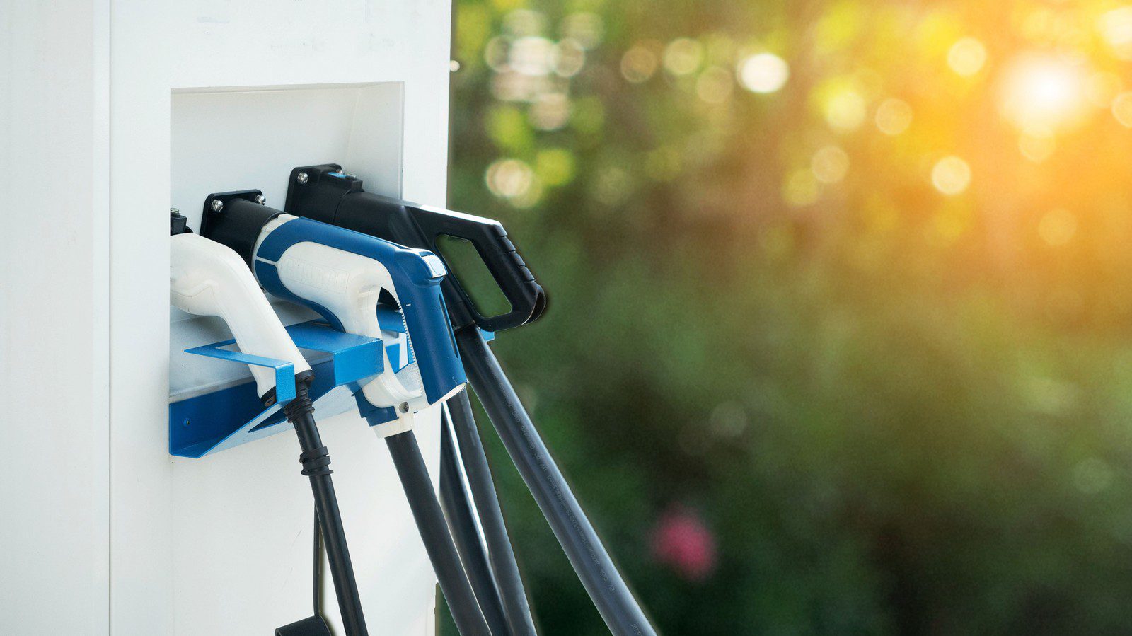 The image shows a charging station for electric vehicles (EVs). There are two EV charging connectors prominently displayed on holders attached to the station, indicating that this is a place where electric cars can be plugged in to recharge their batteries. The background is out of focus with bokeh effects suggesting a bright, sunny day, possibly in a location with greenery.