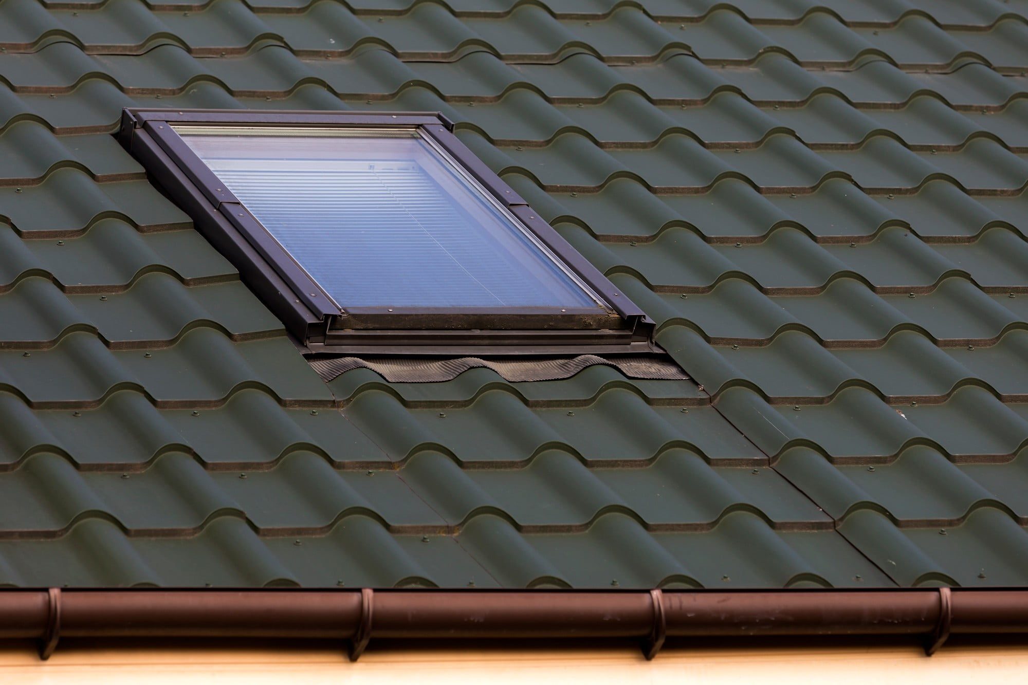 This image shows a tilted rooftop with olive green-coloured roofing tiles. A roof window, often called a skylight, is installed into the slope of the roof, allowing natural light to enter the space below. There's a gutter system with downspouts at the very bottom, indicating the edge of the roof. The materials and design suggest that the building is likely residential and the structure is built to endure various weather conditions while providing functionality and aesthetics.