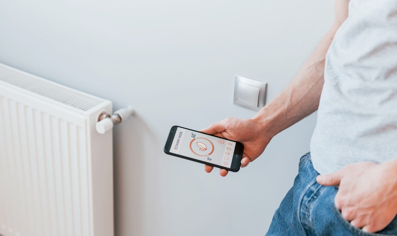 In this image, there is a person holding a smartphone with an app open that appears to be controlling the heating system in a room. The screen of the phone displays an "eco" mode setting and a temperature indicator suggesting energy-saving features. Next to the person is a white radiator, and above this is a wall-mounted thermostat or control panel. The person is casually dressed in a t-shirt and jeans. The focus of the image is on the interaction between smart technology and home heating efficiency.