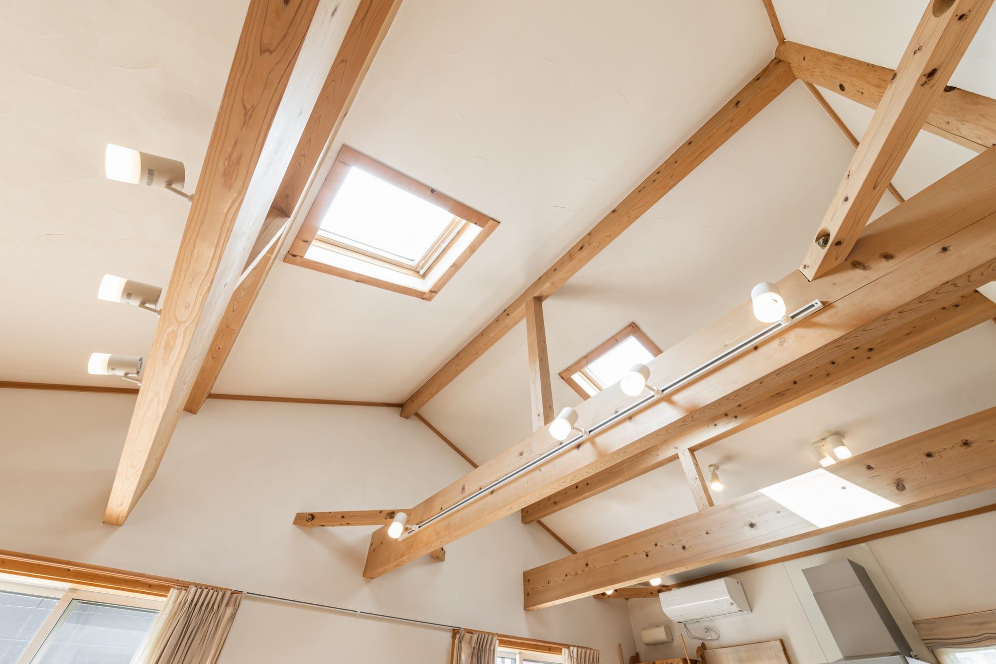 This image shows the interior of a room with a high angled ceiling, which appears to be designed in a contemporary style. The ceiling is adorned with exposed wooden beams that add an aesthetic and structural element to the space. There are track lighting fixtures mounted on some of the beams, each with multiple adjustable light bulbs providing illumination to the room.Additionally, there are two skylights built into the ceiling, allowing natural light to enter and brighten the interior. The walls are painted in a light colour that complements the warmth of the wooden beams. At the peripheral edge of the photo, part of a window with curtains is visible, suggesting the presence of external windows in the room.This setup is characteristic of modern, airy, and well-lit architectural designs often found in residential homes to create an open and inviting space.