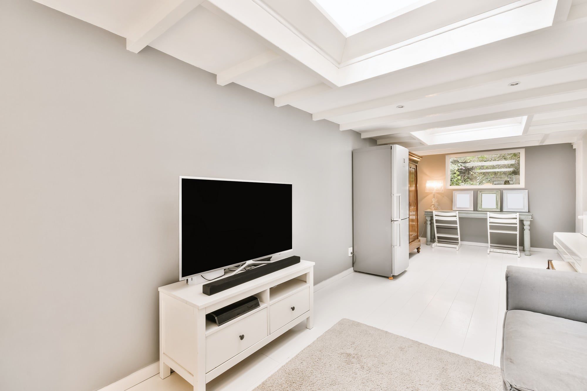 This image shows a modern, well-lit living room. There is white furniture that gives the space a clean and contemporary feel. In the centre, there's a flat-screen TV sitting on a white TV console, which also has a soundbar on it. To the left, we can see the edge of a grey sofa and a small portion of a light-coloured rug.In the background, towards the right, there is a small desk area with two mirrored makeup tables or desks with drawers; above the desks are three framed pictures or mirrors. There's also a large, modern double-door refrigerator in the back right corner. The floors are a light hardwood or laminate, and the room has recessed ceiling lights as well as natural light coming in from a window at the back. The ceiling has distinct architectural elements with inset beams that add character to the space.