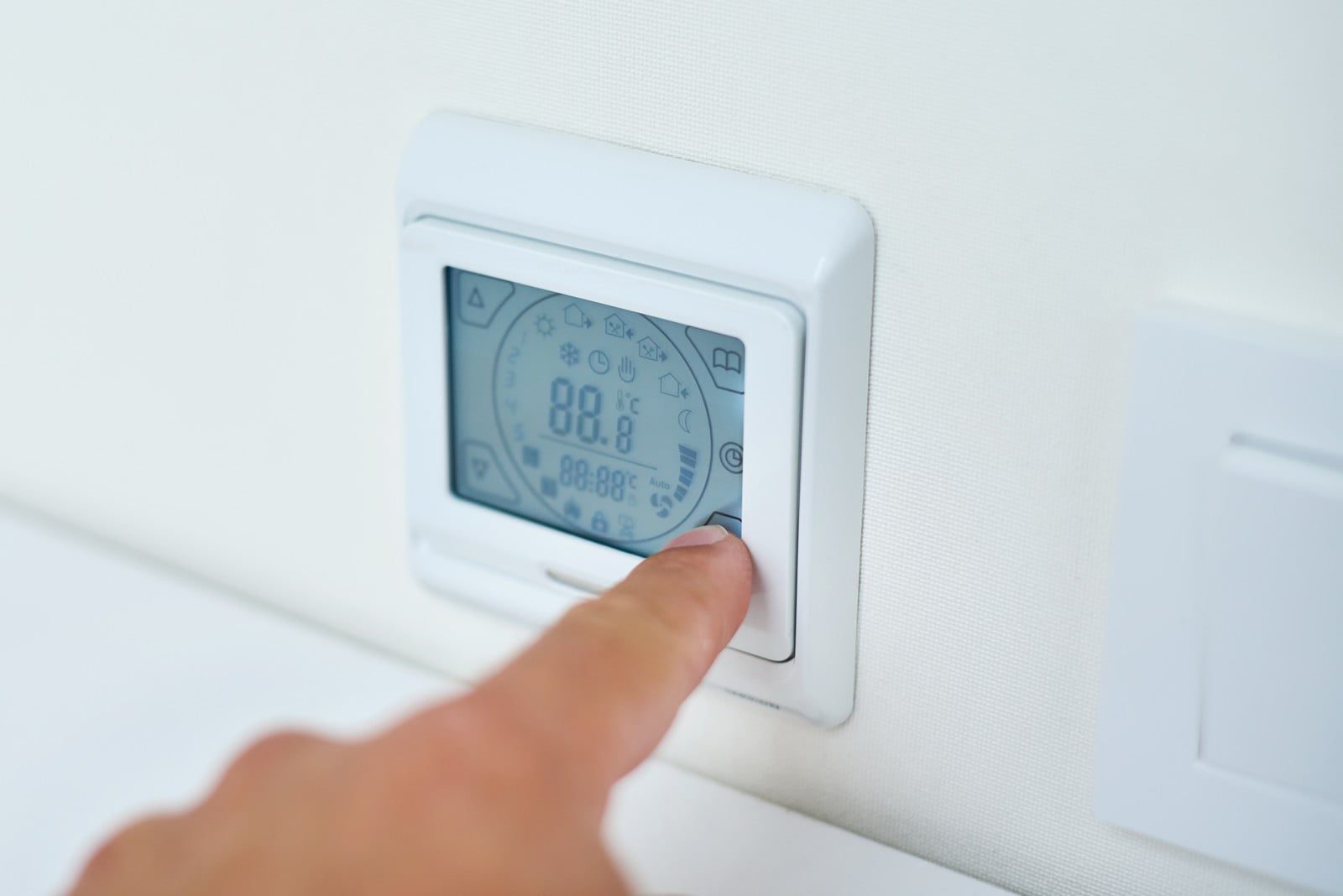 The image displays a programmable digital thermostat mounted on a wall. A person's hand is pressing one of the buttons, presumably to adjust the settings. Thermostats like this are used to control the temperature of a room or building by turning a heating or cooling system on or off, or by regulating the flow of a heat carrier. The digital display indicates the temperature and may show other configurable settings like time, operating mode, and other parameters.