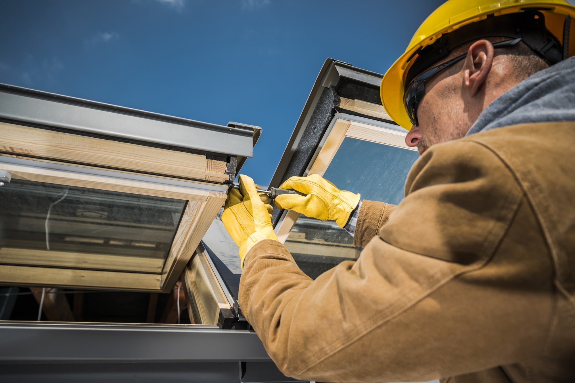 The image shows a person engaged in construction or maintenance work, specifically working on a window. The individual is wearing a yellow hard hat, safety glasses, and yellow work gloves, implying adherence to safety protocols typically observed at a construction site. This person appears to be installing or repairing a window frame, as they are holding onto the frame or adjusting something related to the window's casing or hardware. The window has multiple panes and is hinged at the top. It seems to be part of a larger structure, possibly a residential or commercial building.The setting is outdoors, with a clear blue sky in the background, suggesting a fair weather condition conducive to outdoor work. The focus of the image is on the worker's hands and the immediate task, with the worker's identity obscured as we mostly see the side and back of their head. There is an emphasis on the action and the details of the labour rather than the individual performing it.