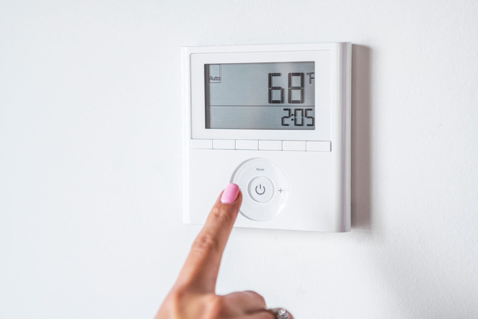 The image shows a digital thermostat mounted on a white wall. A person's finger, with a pink nail, is pressing the power button of the thermostat. The display of the thermostat indicates a temperature of 68 degrees Fahrenheit and a time of 2:05. The thermostat also has additional control buttons and a mode selection area, indicating its programmability for controlling the temperature within the space it is installed in.