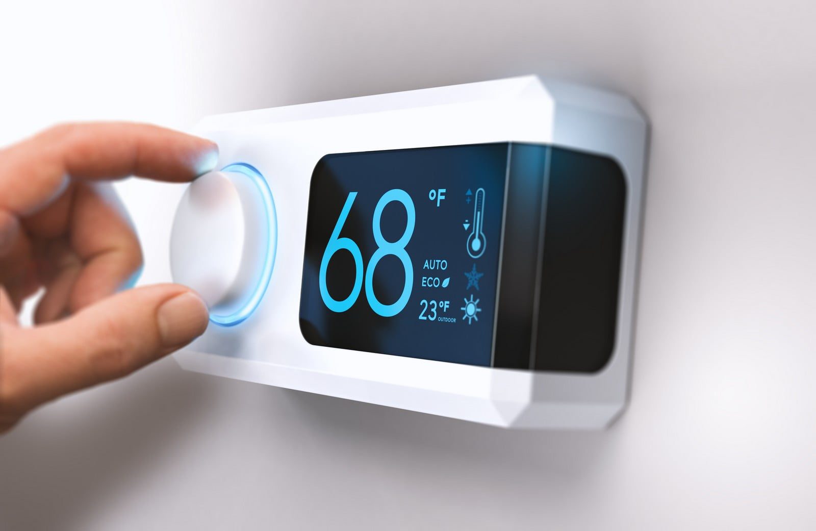 The image shows a close-up of a person's hand adjusting a modern digital thermostat on a wall. The thermostat has a sleek design with a touchscreen display showing a temperature of 68°F. There are additional settings visible on the screen, such as "AUTO" mode and an "ECO" function, along with an outdoor temperature reading of 23°F, indicated by a small snowflake icon. The person is turning a dial, which is illuminated with a blue light indicating interaction. This type of thermostat is typically used for regulating heating and cooling systems in a building or home for comfort and energy efficiency.