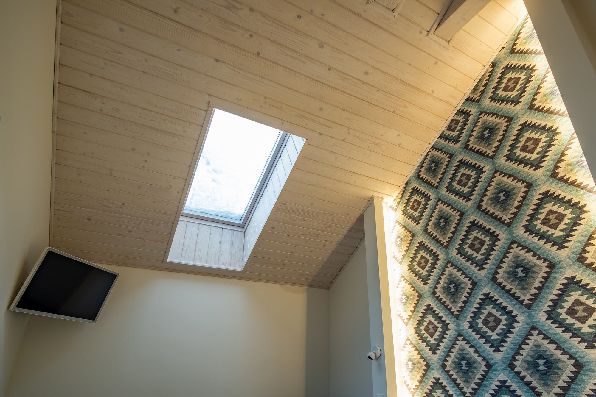 The image shows a room with a vaulted ceiling paneled with light wood. A skylight is installed in the sloped part of the ceiling, allowing natural light to filter into the space. On the left-hand side, there's a flat-screen TV mounted on the wall. The opposite wall presents a patterned textile or rug hanging as a decorative piece. The colour and pattern give the room a cosy and possibly eclectic aesthetic. It appears to be an interior of a modern home with a focus on utilising natural light and combining functional design with decorative elements.